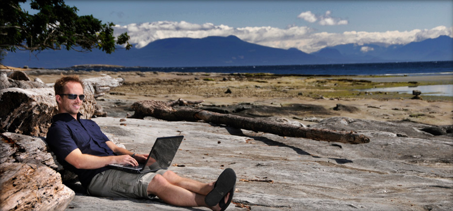 A man sits on a hard rock beach with a laptop near the ocean